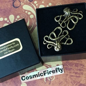 May include: Two bronze-colored octopus-shaped cufflinks presented in a black box. The cufflinks have detailed tentacles and a textured surface. The box is open, revealing the cufflinks against a patterned background. The box has a sticker that says "Handcrafted By CosmicFirefly.com Las Vegas".
