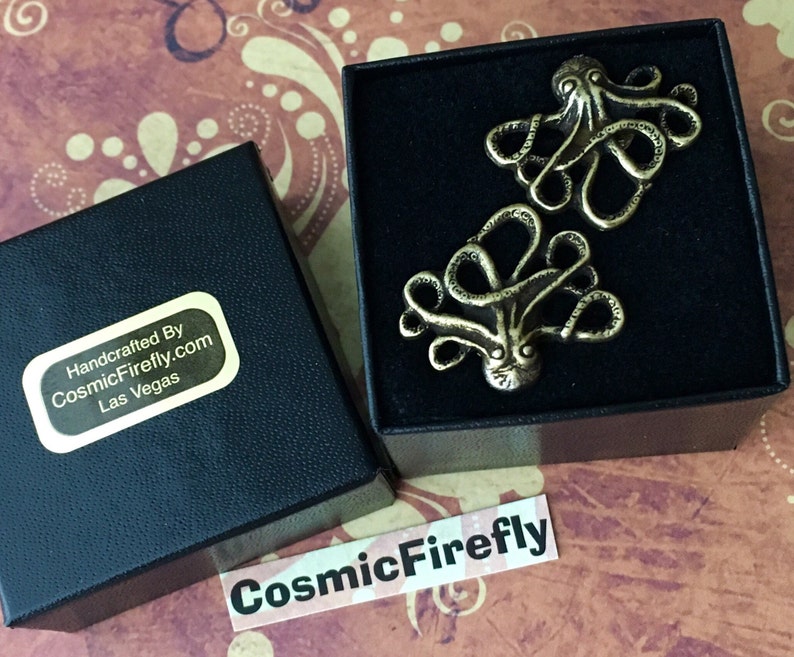 May include: Two bronze-colored octopus-shaped cufflinks presented in a black box. The cufflinks have detailed tentacles and a textured surface. The box is open, revealing the cufflinks against a patterned background. The box has a sticker that says "Handcrafted By CosmicFirefly.com Las Vegas".