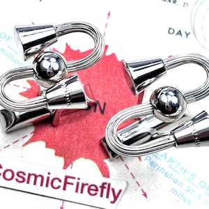 May include: Silver cufflinks with a distinctive design. Each cufflink has an S-shaped bar with a central sphere and flared ends. The background shows a map and the text "CosmicFirefly". The cufflinks are likely made of a shiny metal.