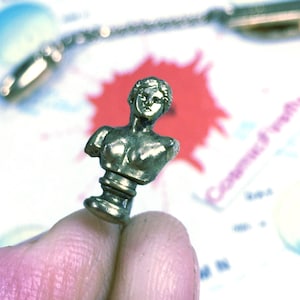 May include: A small, antique-style brass bust of a classical figure. The bust is detailed with a sculpted face, shoulders, and chest. The figure is mounted on a decorative base. The background includes a blurred spoon and a red and white background.