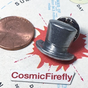 May include: A silver-colored metal top hat lapel pin positioned next to a copper penny for scale. The pin features a classic cylindrical crown and a wide brim. Below the pin is a white rectangular label with the text CosmicFirefly in red.