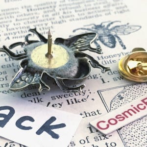 May include: A silver-toned bee-shaped lapel pin shown from the back, featuring a central metal post and a separate gold-toned butterfly clutch backing. A white paper label with the black text Back and a label with CosmicFirefly are visible.