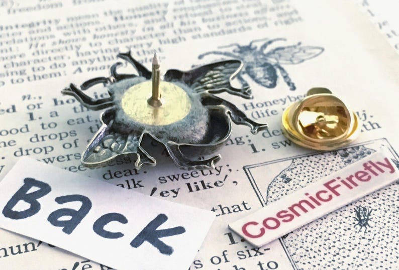 May include: A silver-toned bee-shaped lapel pin shown from the back, featuring a central metal post and a separate gold-toned butterfly clutch backing. A white paper label with the black text Back and a label with CosmicFirefly are visible.