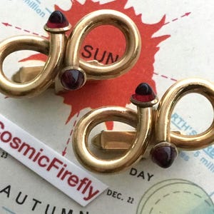 May include: A pair of gold-toned cufflinks with a unique design. Each cufflink features a curved shape with a red gemstone accent at the top and bottom. The cufflinks are displayed on a vintage map with the text "CosmicFirefly".