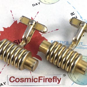 May include: Two gold-coloured cufflinks with a ribbed barrel design. The cufflinks have a rectangular bar and a cylindrical body with horizontal ridges. The background features a map with the text "CosmicFirefly".