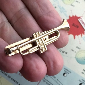 Peut inclure: Une pince à cravate en forme de trompette dorée. Le petit instrument de musique est très détaillé, avec des pistons et un pavillon visibles. La pince est conçue pour être fixée à une cravate, ajoutant une touche musicale à une tenue formelle. La trompette mesure environ 5 cm de long.