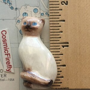 May include: A small ceramic Siamese cat figurine with blue eyes and brown markings. The cat is sitting with its tail curled. The figurine is approximately 3.8 cm tall. The CosmicFirefly label is visible in the background.