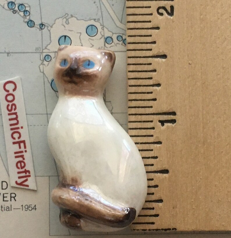 May include: A small ceramic Siamese cat figurine with blue eyes and brown markings. The cat is sitting with its tail curled. The figurine is approximately 3.8 cm tall. The CosmicFirefly label is visible in the background.