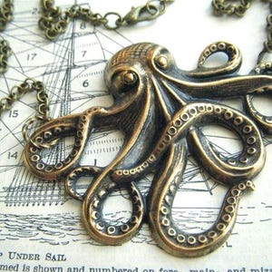 May include: Bronze-toned octopus pendant necklace. The detailed pendant features an octopus with textured tentacles and suction cups. The necklace chain is also bronze-toned. The pendant rests on a vintage nautical chart.