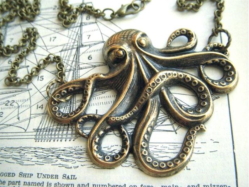 May include: Bronze-toned octopus pendant necklace. The detailed pendant features an octopus with textured tentacles and suction cups. The necklace chain is also bronze-toned. The pendant rests on a vintage nautical chart.