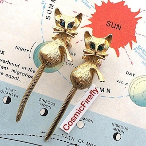 May include: Two gold-toned cat-shaped brooches with blue gemstone eyes. The cats have textured bodies and long, slender tails. The brooches are displayed on a vintage map with the text "CosmicFirefly".