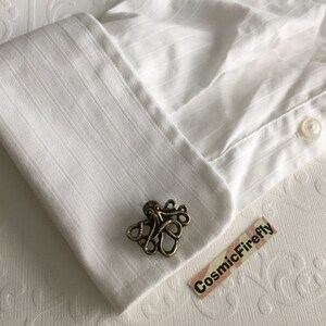 May include: A bronze-colored octopus cufflink rests on a white dress shirt cuff. The cufflink is detailed with tentacles and a central body. The shirt has vertical stripes and a button closure. The background is a textured white surface.