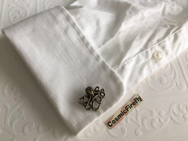 May include: A bronze-colored octopus cufflink rests on a white dress shirt cuff. The cufflink is detailed with tentacles and a central body. The shirt has vertical stripes and a button closure. The background is a textured white surface.