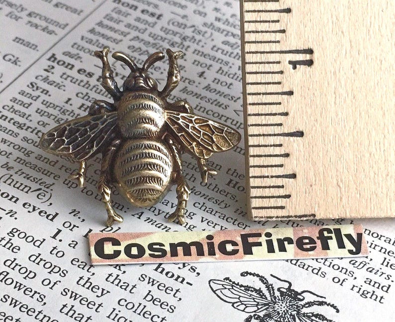 May include: A brass-toned bee-shaped pin featuring detailed wings and body segments, positioned next to a ruler showing a width of approximately 1.25 inches. A small label with the text CosmicFirefly is placed below the bee pin.
