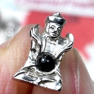 May include: A silver-coloured figurine of a seated figure wearing a hat, possibly a deity or spiritual figure. The figurine has a black, round stone set in the centre. The figure is held in a person's hand.