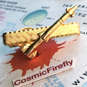 Könnte beinhalten: Eine goldfarbene Krawattenklammer mit Raketendesign. Die Klammer hat eine strukturierte rechteckige Basis und ein detailliertes Raketenmodell. Der Text "CosmicFirefly" ist auf einem kleinen weißen Etikett zu sehen.