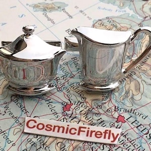 May include: A silver-plated sugar bowl with a lid and a creamer set on a map. The sugar bowl has a lid with a small handle. The creamer has a handle and a spout. The set is reflective and shiny.
