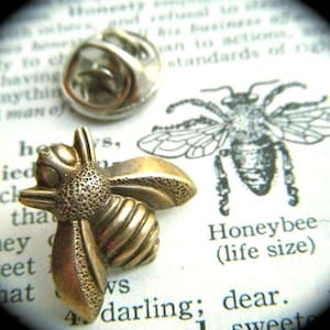 May include: A brass-toned metal bee lapel pin featuring a textured body and wings, shown with its silver-toned clutch back. The pin is placed on a page of text that includes the printed words Honeybee (life size) and a line drawing of a bee.