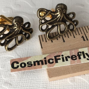 May include: Bronze-colored octopus cufflinks with gold-colored hardware. The cufflinks feature a detailed octopus design. The text "CosmicFirefly" is printed on a small label.
