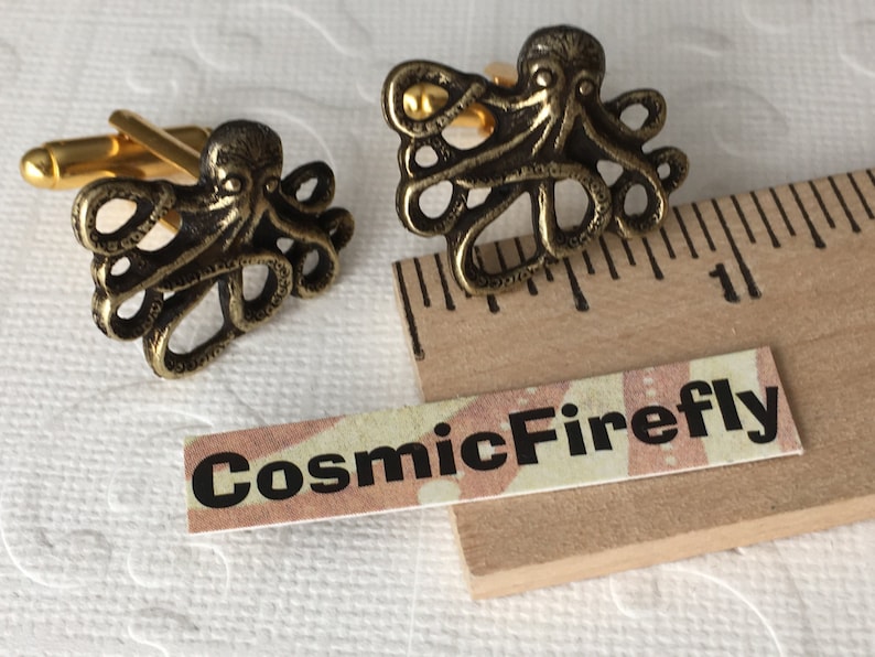 May include: Bronze-colored octopus cufflinks with gold-colored hardware. The cufflinks feature a detailed octopus design. The text "CosmicFirefly" is printed on a small label.