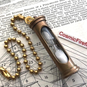 May include: A vintage-style hourglass pendant with a brass-colored metal casing and a chain of small, round beads. The hourglass contains white sand and is suspended against a backdrop of nautical charts. The pendant has the text "CosmicFirefly".