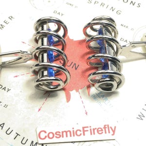 May include: A pair of silver-coloured cufflinks with a distinctive spiral design. Each cufflink has a central blue accent. The cufflinks are presented on a vintage map background.