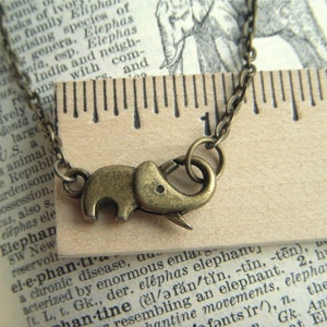 May include: A bronze-toned elephant pendant necklace. The elephant charm is detailed with a trunk and small tusks. The necklace chain is also bronze-toned. The background includes a ruler and text from a dictionary page.