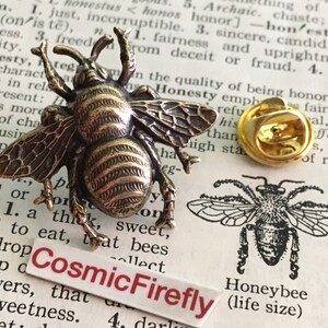 May include: A bronze-toned bee lapel pin with detailed wings and body texture, accompanied by a separate gold-colored metal clutch back. A small white rectangular label with the red text CosmicFirefly is positioned below the bee.