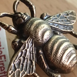 May include: A bronze-toned bee figurine with detailed wings and a textured body. A white rectangular tag with the red text CosmicFirefly is positioned next to the insect, which features prominent antennae and legs.