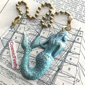 May include: A blue mermaid pendant with a brass chain and clasp. The mermaid has a textured finish and is lying down. The pendant is attached to a brass ball chain. The text "CosmicFirefly" is on a white label.