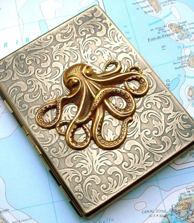 Op de afbeelding: Een rechthoekige koffer in antieke stijl met een sierlijk, gegraveerd bloemmotief. Een goudkleurige octopus is op de bovenkant bevestigd. De koffer heeft een gedempte goudkleur en de achtergrond is een kaart.