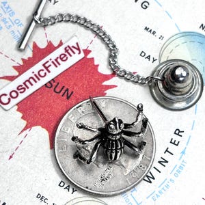 Könnte beinhalten: Ein silberfarbener Krawattenstecker mit detailliertem Insektenmuster, befestigt an einer Kette und einem weißen rechteckigen Anhänger mit dem Text "CosmicFirefly". Der Stecker befindet sich auf einem Hintergrund im Kartenstil.