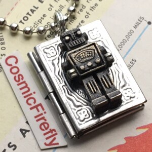May include: A silver-toned locket pendant with a detailed robot charm. The robot is dark bronze and sits atop a silver book-shaped locket. The pendant hangs from a silver ball chain. The text "CosmicFirefly" is visible on a white label.