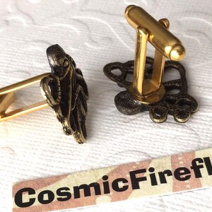 May include: A pair of gold-toned cufflinks with an octopus design. The octopus is dark bronze in color. The cufflinks have a classic bar and toggle closure. The brand name "CosmicFirefly" is printed on a small label.