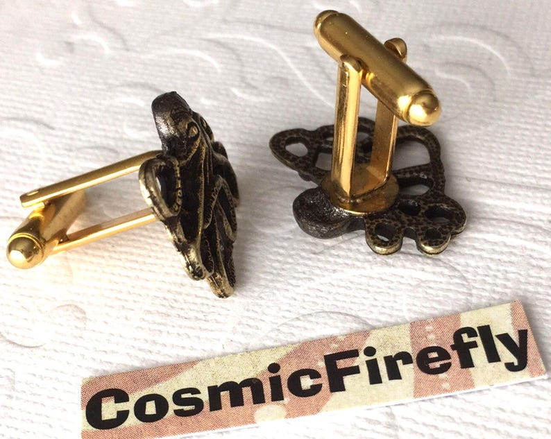 May include: A pair of gold-toned cufflinks with an octopus design. The octopus is dark bronze in color. The cufflinks have a classic bar and toggle closure. The brand name "CosmicFirefly" is printed on a small label.