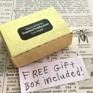 May include: A rectangular, pale yellow gift box with a textured surface and a black label reading Handcrafted by CosmicFirefly.com Las Vegas. A white paper note with the handwritten black text FREE GIFT Box included is placed below.