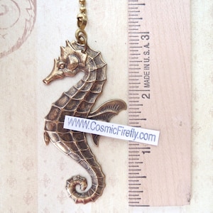 May include: A bronze-colored seahorse pendant with intricate detailing, suspended from a gold-tone chain. The seahorse is approximately 2 inches in length. The pendant has a textured surface and a curled tail. The text "WWW.CosmicFirefly.com" is visible.
