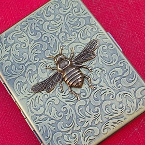 K&ouml;nnte beinhalten: Ein Zigarettenetui aus Messing mit einem verzierten Blumenmuster und einer Bienenverzierung.