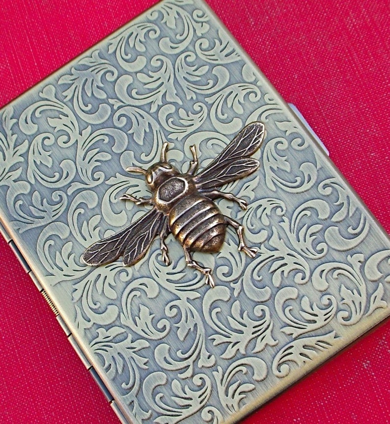 K&ouml;nnte beinhalten: Ein Zigarettenetui aus Messing mit einem verzierten Blumenmuster und einer Bienenverzierung.