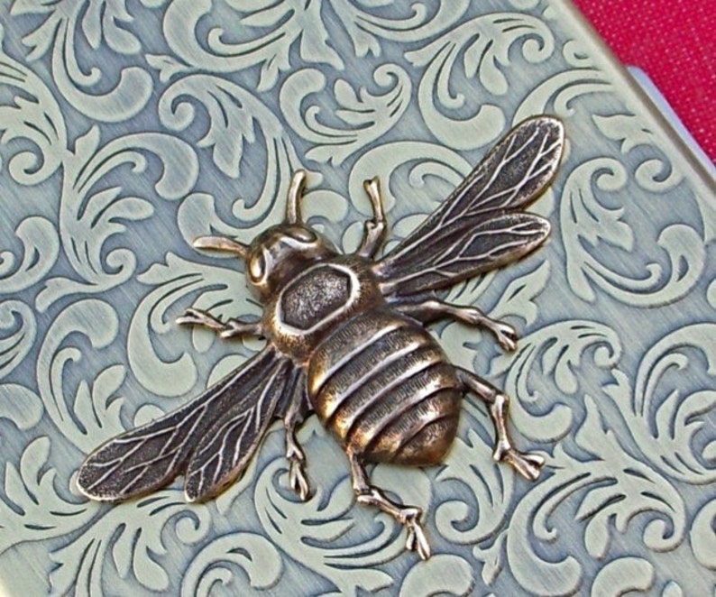 K&ouml;nnte beinhalten: Eine detailreiche, verzierte, messingfarbene Biene mit einer strukturierten, metallischen Oberfl&auml;che. Die Biene ist vor einem Hintergrund aus einem dekorativen, gepr&auml;gten Blumenmuster platziert.