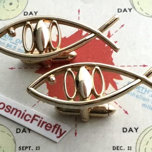 May include: Gold-coloured cufflinks in a fish shape. Each cufflink has an open frame with three oval cut-outs and a central, elongated oval. The cufflinks are displayed on a vintage map, with the text "CosmicFirefly".