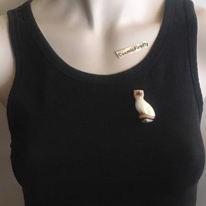 May include: A cream and brown Siamese cat brooch. The cat is depicted in a seated position with blue eyes. The brooch is attached to a black vest top. The text "CosmicFirefly" is on a small label.