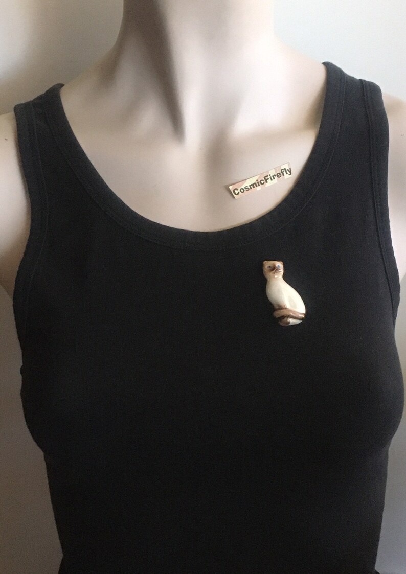 May include: A cream and brown Siamese cat brooch. The cat is depicted in a seated position with blue eyes. The brooch is attached to a black vest top. The text "CosmicFirefly" is on a small label.