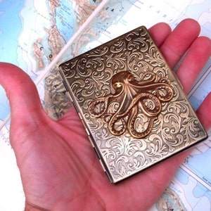 Op de afbeelding: Een rechthoekige metalen koffer in antieke stijl met een reli&euml;f bloemmotief en een bronzen octopus embleem. De koffer wordt in een hand gehouden, met een kaart zichtbaar op de achtergrond. De koffer is ongeveer 9 cm lang.