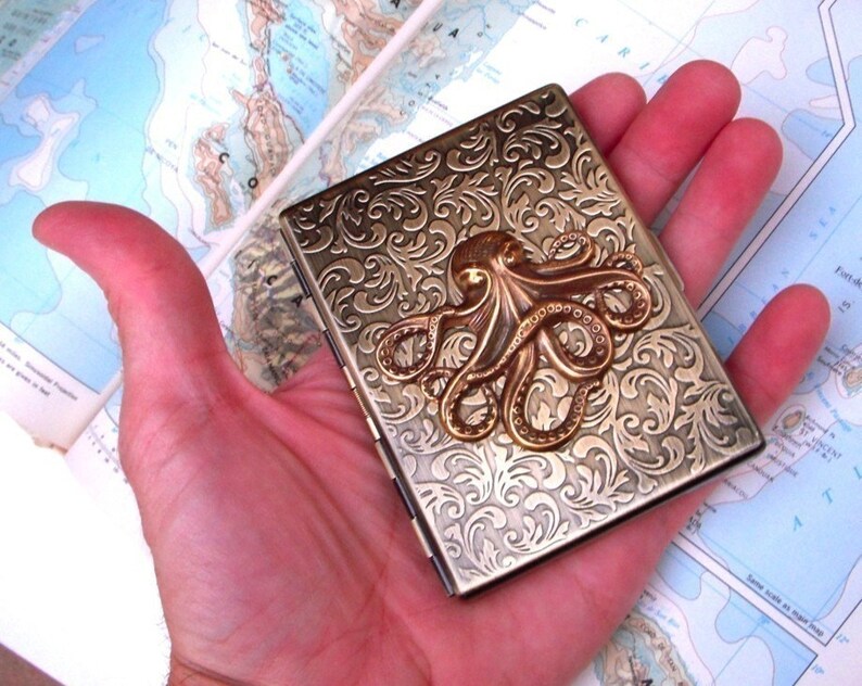 Op de afbeelding: Een rechthoekige metalen koffer in antieke stijl met een reli&euml;f bloemmotief en een bronzen octopus embleem. De koffer wordt in een hand gehouden, met een kaart zichtbaar op de achtergrond. De koffer is ongeveer 9 cm lang.