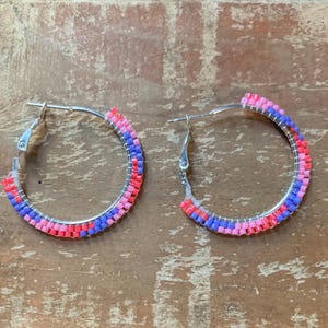 May include: A pair of silver hoop earrings with a beaded design. The beads are arranged in bands of red, pink, and blue. The earrings are displayed on a wooden surface.