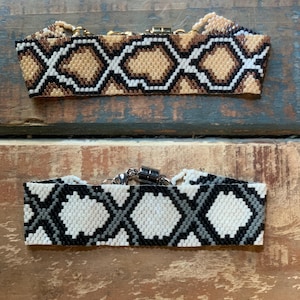 Peut inclure: Deux colliers ras du cou en perles avec un motif géométrique. Le collier du haut est brun, blanc et noir. Le collier du bas est blanc, noir et gris.