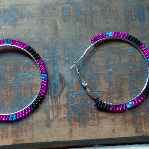 May include: A pair of silver hoop earrings with a beaded design. The beads are arranged in bands of black, magenta, and blue. The earrings are displayed on a weathered, blue-toned wooden surface.