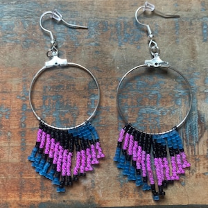 May include: A pair of hoop earrings with a beaded fringe. The earrings have silver-tone hoops and a fringe of blue, black, and purple beads. They are displayed on a rustic wooden surface.