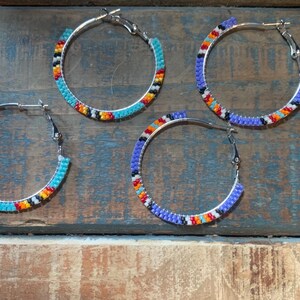May include: Four silver hoop earrings with colourful beaded designs. The earrings feature turquoise, orange, yellow, black, and purple beads. The earrings are displayed on a rustic, weathered wooden surface.